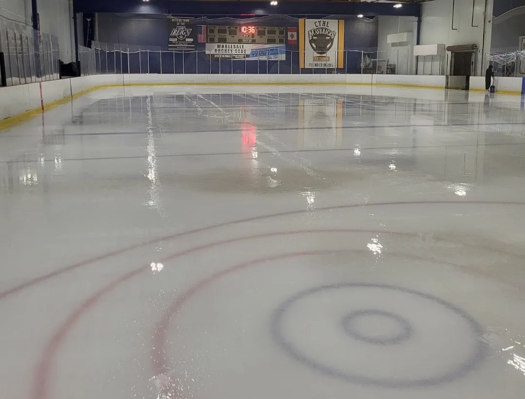 Hielo recién pulido y vacío en Ice Realm Carlsbad, con un círculo de saque al frente y el marcador y las mantas de los equipos bajo un techo de madera — Ice Realm Carlsbad