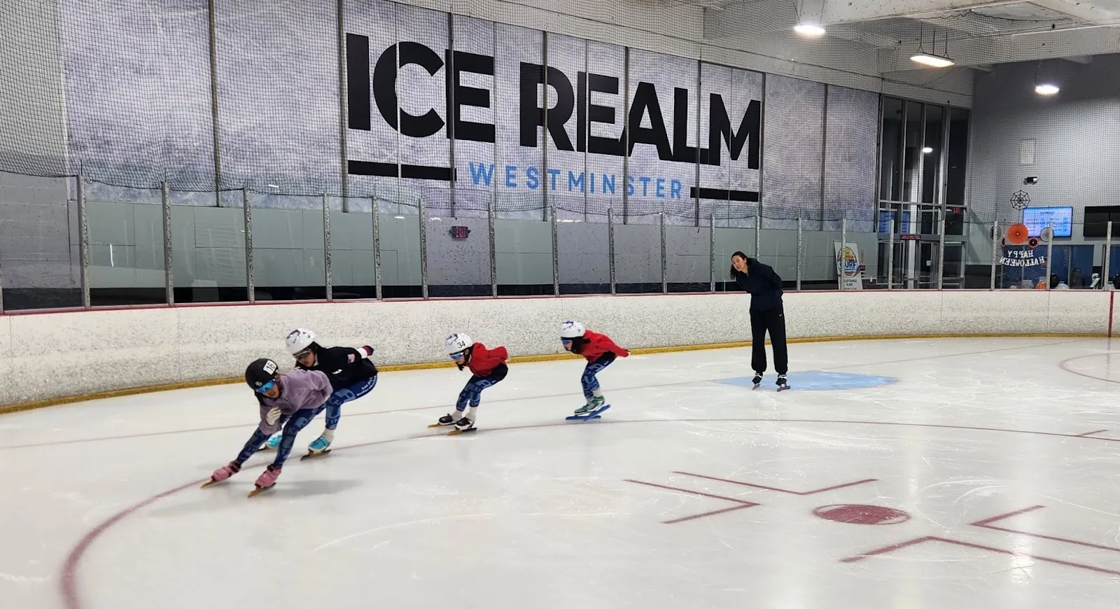 Patinadores inclinándose en una curva a lo largo de las vallas del fondo, con el nombre Ice Realm en letras grandes sobre la pared de vidrio detrás de una amplia pista blanca. — Ice Realm Westminster
