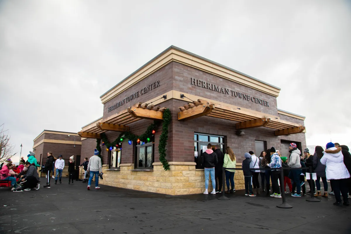 The Herriman Towne Center building beside the ice ribbon, with visitors queuing at its windows under a pergola roof hung with garland. — Ice Ribbon at J. Lynn Crane Plaza