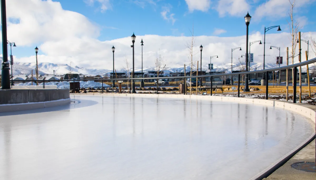 The curved ice ribbon at J. Lynn Crane Plaza in Herriman on a bright winter day, with a rail, street lamps and snowy mountains beyond. — Ice Ribbon at J. Lynn Crane Plaza