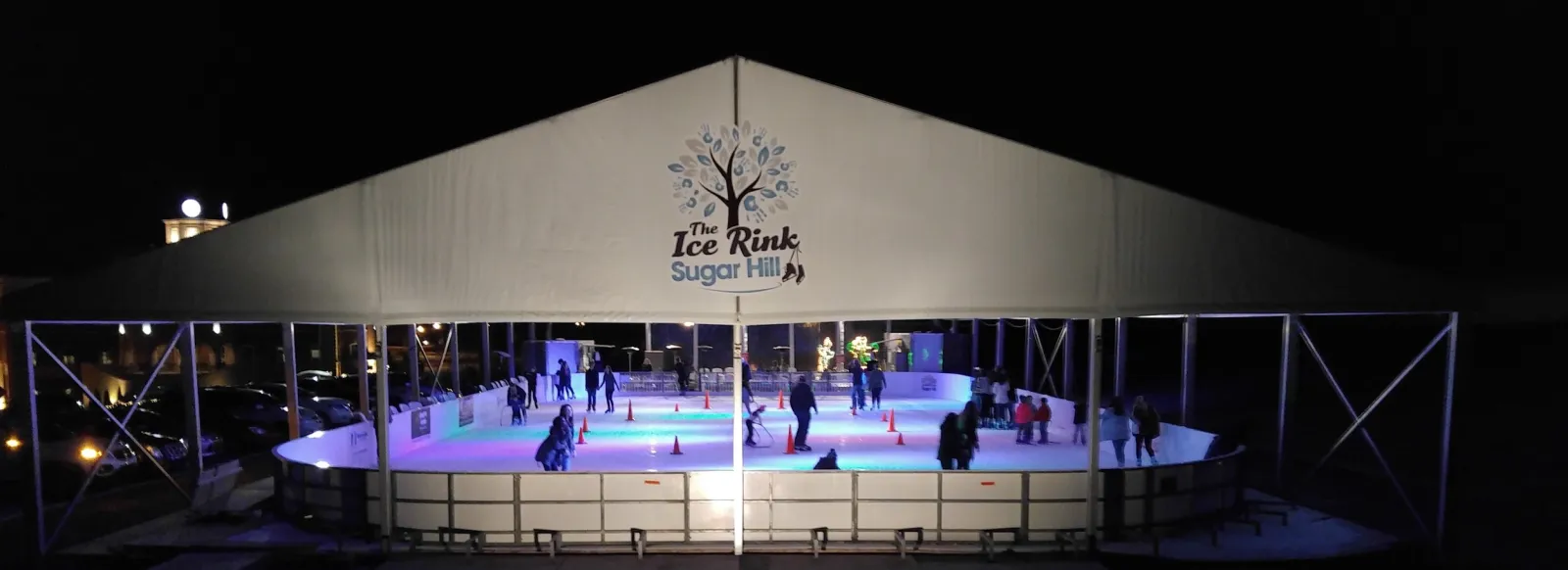 The Ice Rink Sugar Hill under its white peaked tent at night, the tree-and-skates logo on the gable and skaters on the lit ice behind the boards. — The Ice Rink at Sugar Hill