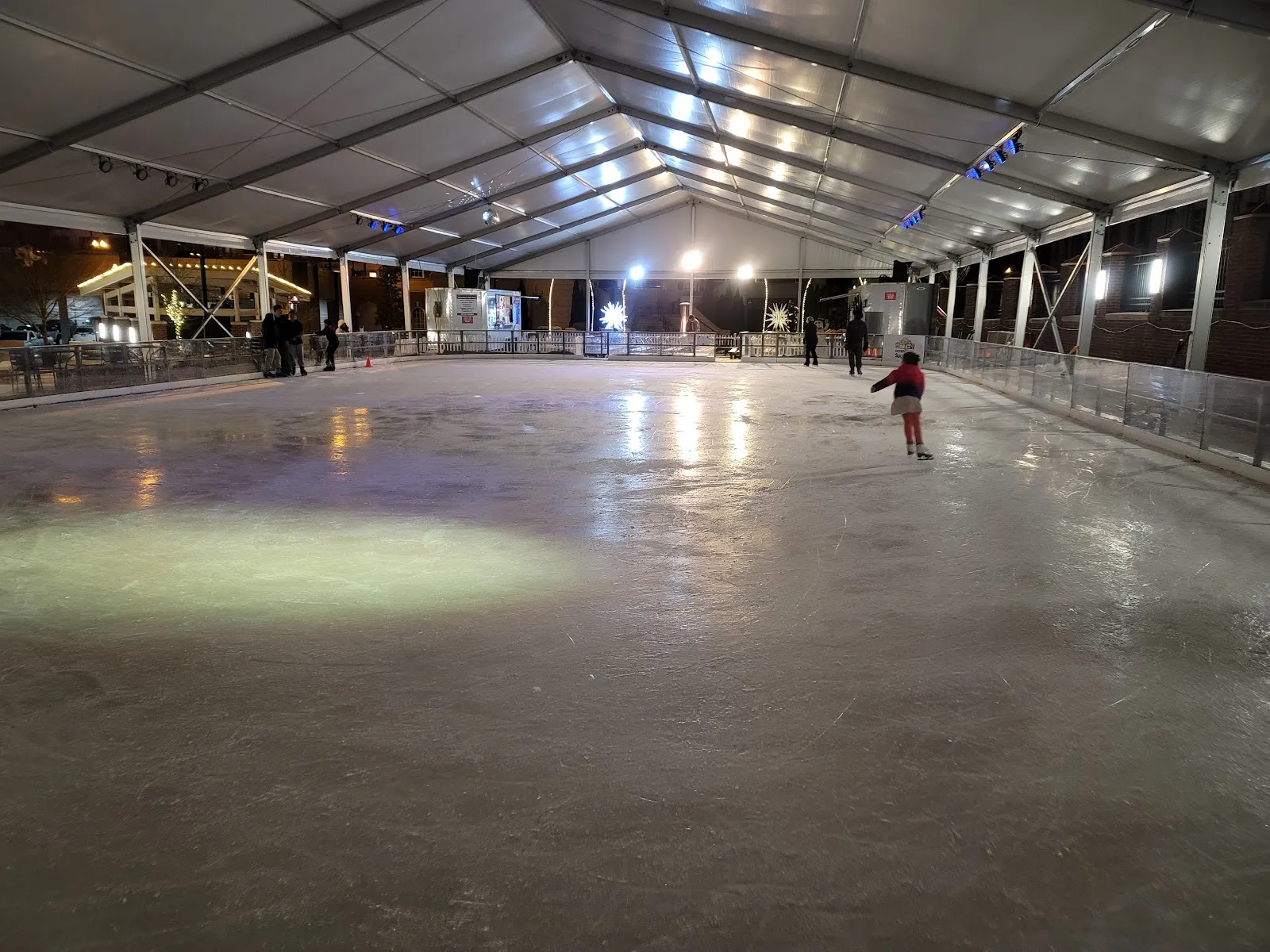 The full sheet of ice under the rink tent at night, lit by floodlights, with a few skaters at the far end. — The Ice Rink at Sugar Hill