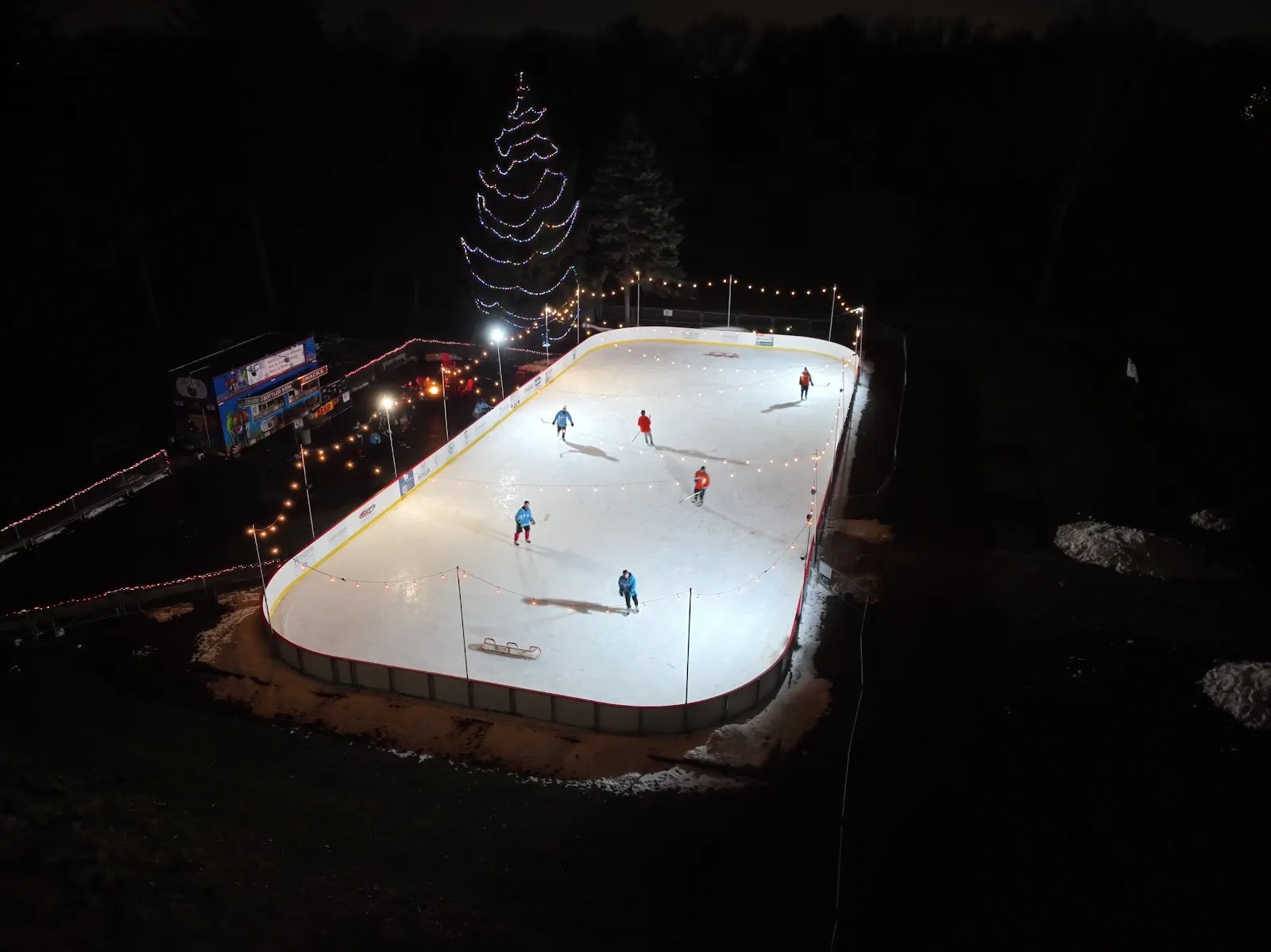 Toda la pista iluminada de noche desde arriba, con patinadores repartidos por el hielo y tiras de luces alrededor de las bandas. — The Ice Rink at Holliday Park