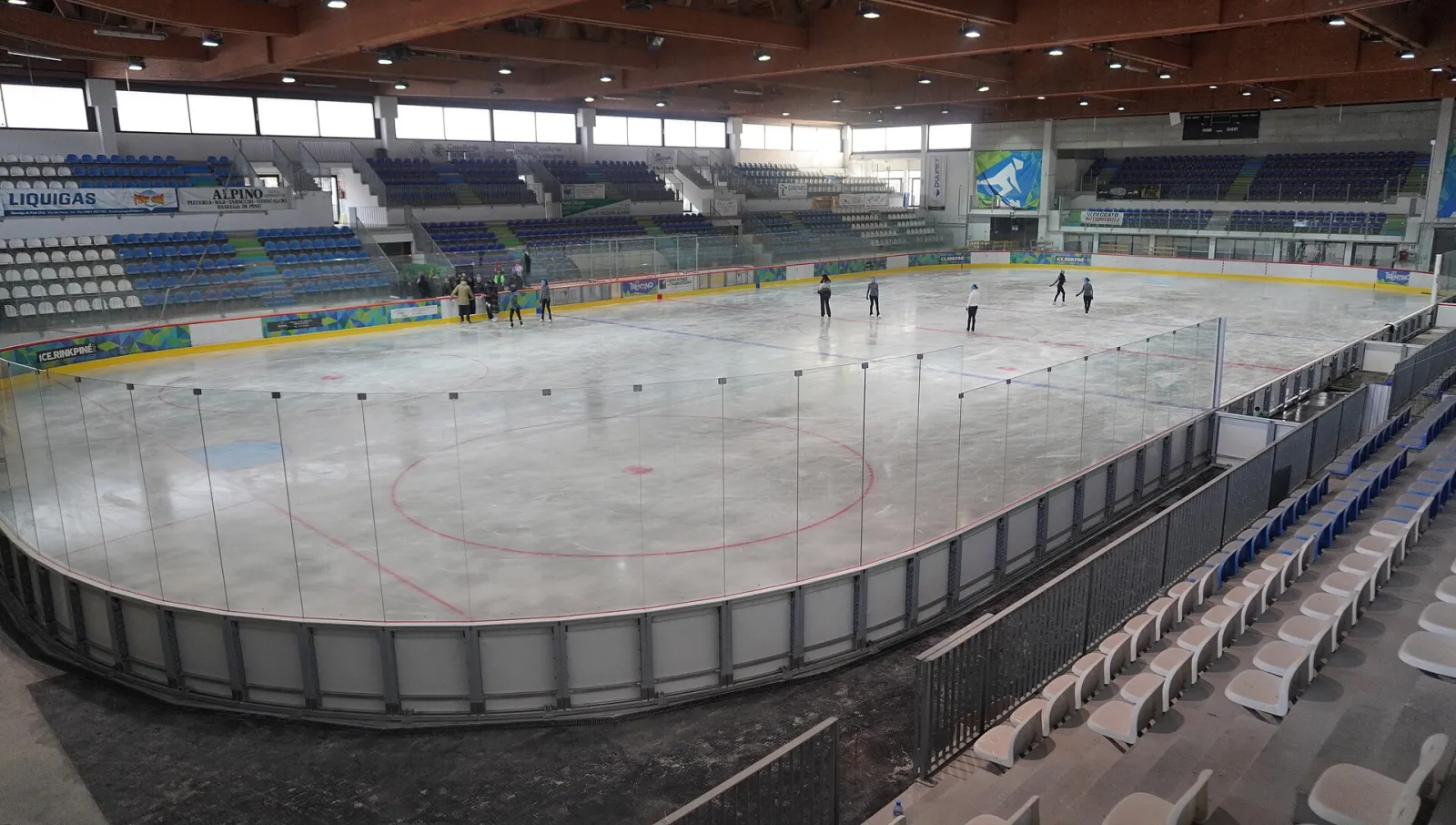Inside the Ice Rink Piné hall: the 60 by 30 metre ice sheet with a few skaters, the stands in blue and white and the timber roof. – Ice Rink Piné
