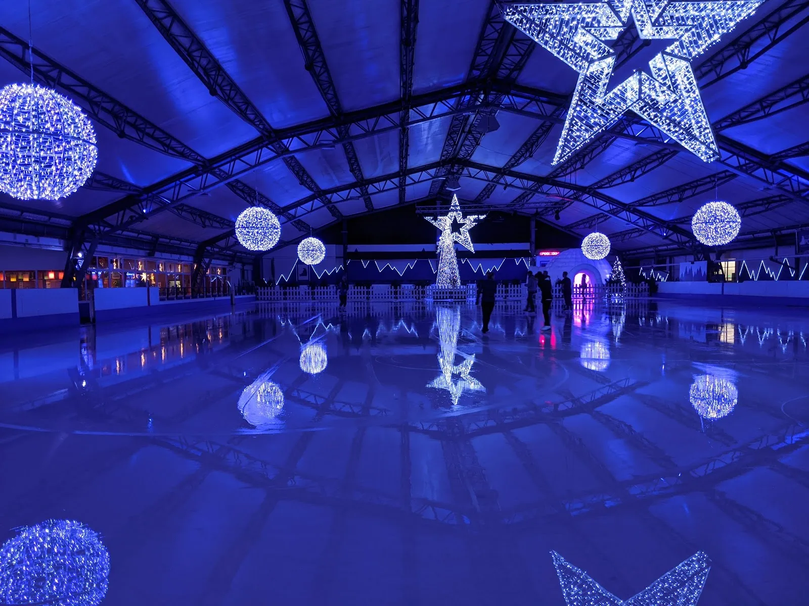 La glace vide de la Patinoire Poseidon, décorée pour l’hiver, éclairée en bleu sous la toiture à charpente d’acier, avec des étoiles et des sphères scintillantes qui se reflètent dans la glace — Ice Skating Poseidon