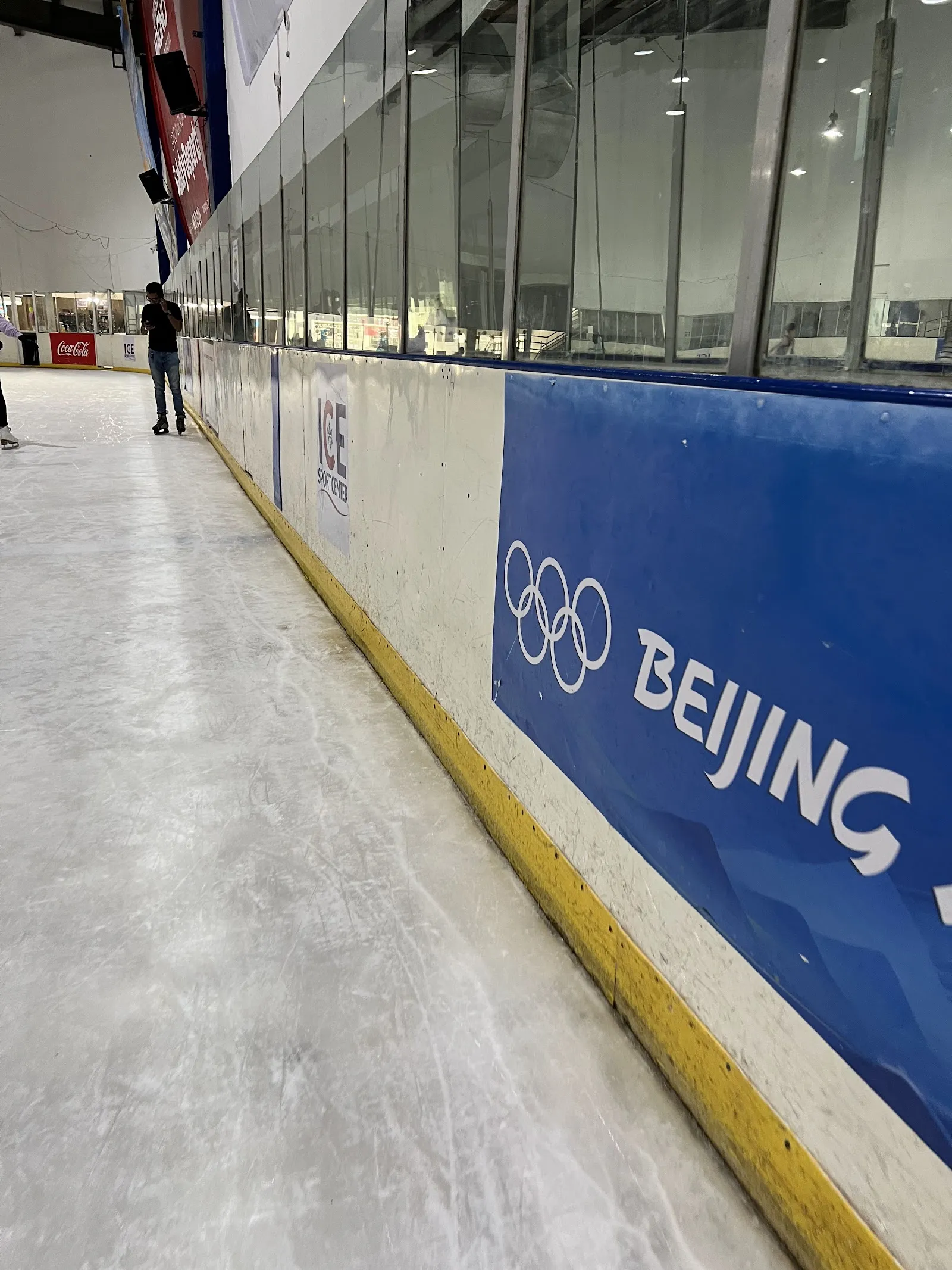 The ice along the boards at Ice Sport Center, with the glass above the dasher and a blue Beijing banner, and one person far down the sheet — Ice Sport Center