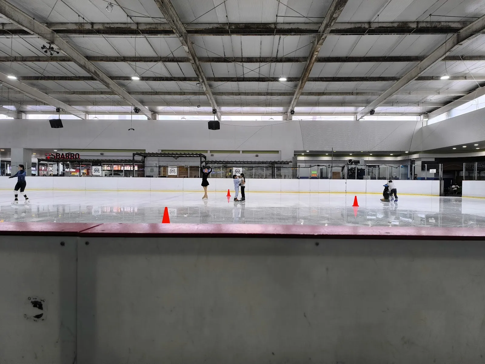 The ice sheet inside the Forum Buenavista mall seen from the boards, with a few skaters and cones far across the ice under the steel ceiling — Ice Station Buenavista
