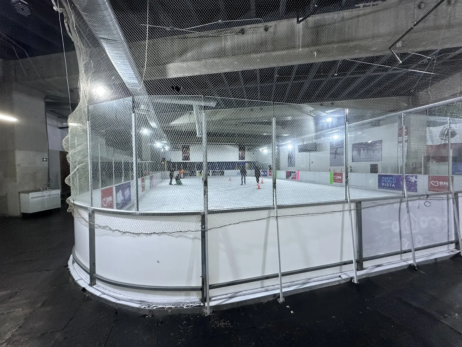 The boarded rink at Ice Town in the Sentura mall seen from outside the netting, with a few skaters far across the ice — Ice Town Sentura