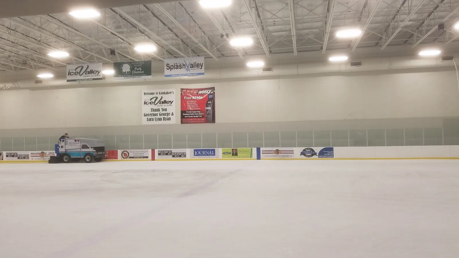 The empty ice seen from rink level, a resurfacer at the far boards beneath Ice Valley Centre and Kankakee Valley Park District banners. — Ice Valley Centre