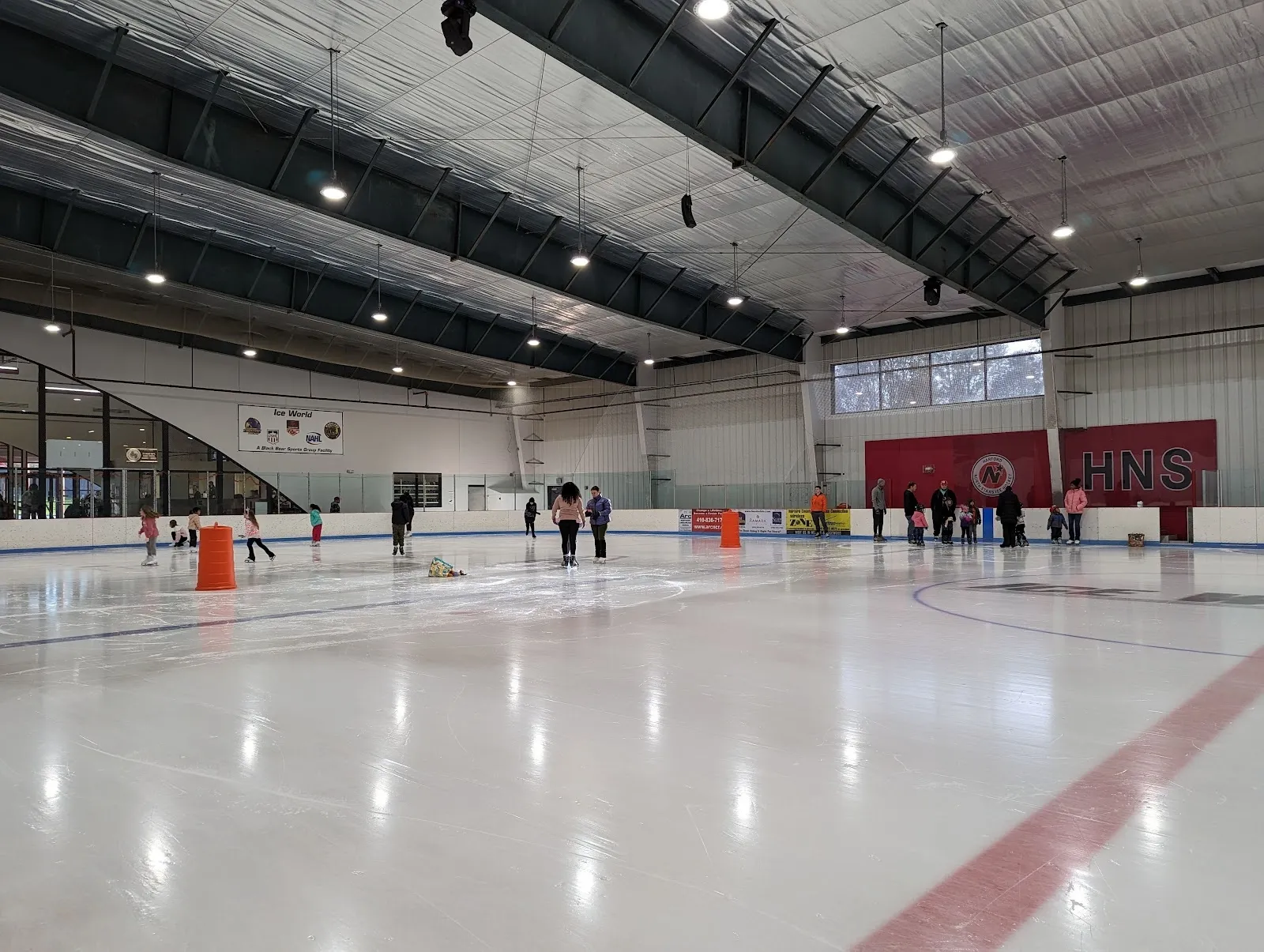 Una pista amplia en una sesión pública, con patinadores repartidos por el hielo entre conos naranjas bajo un largo techo con refuerzos oscuros y banderines rojos en el extremo opuesto. — Ice World