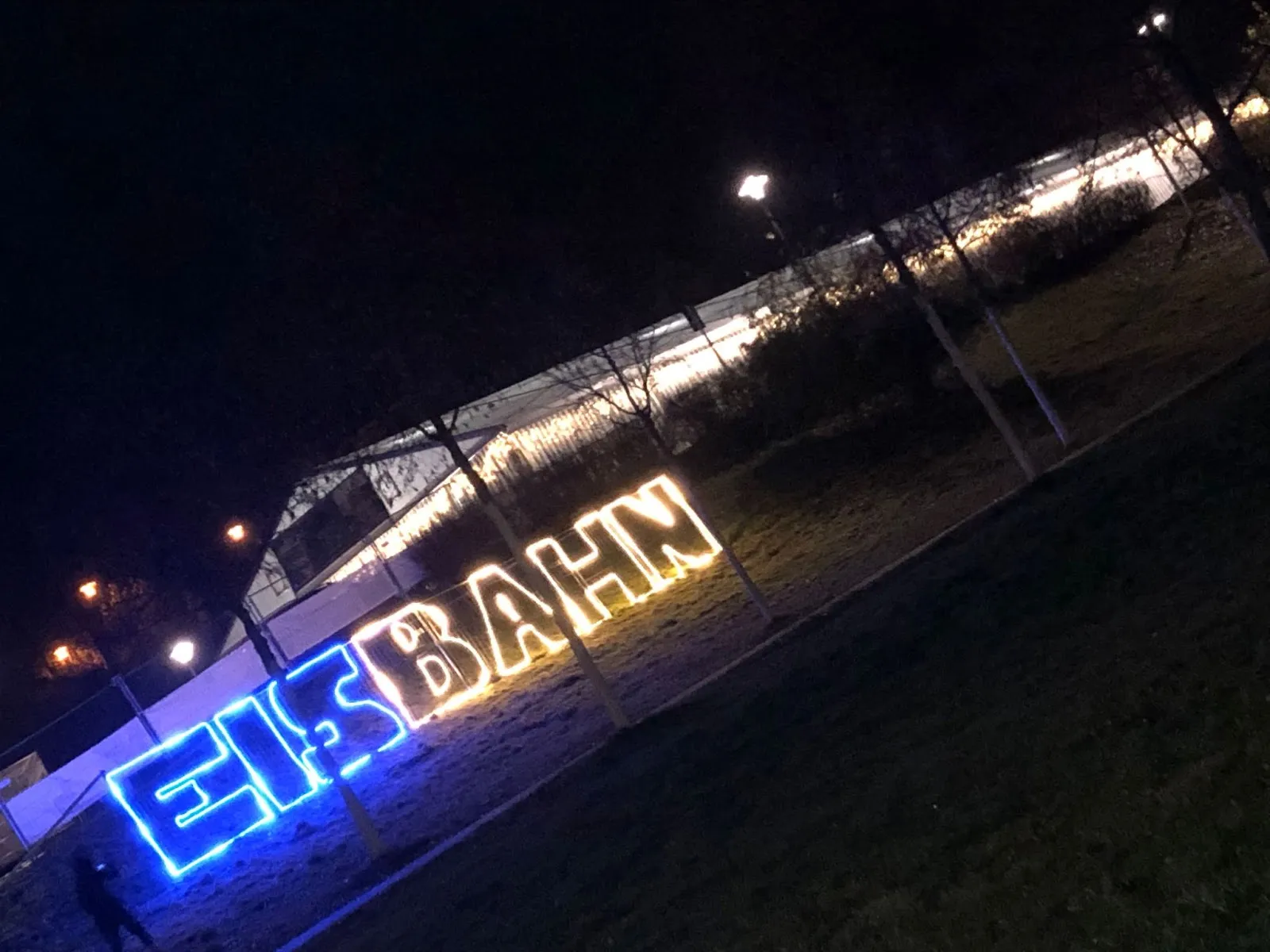 The tent hall at night, lit from within along its roofline, with a glowing EISBAHN sign in blue and yellow on the park lawn in front. — Ice4Moor Eisbahn Bad Schwalbach