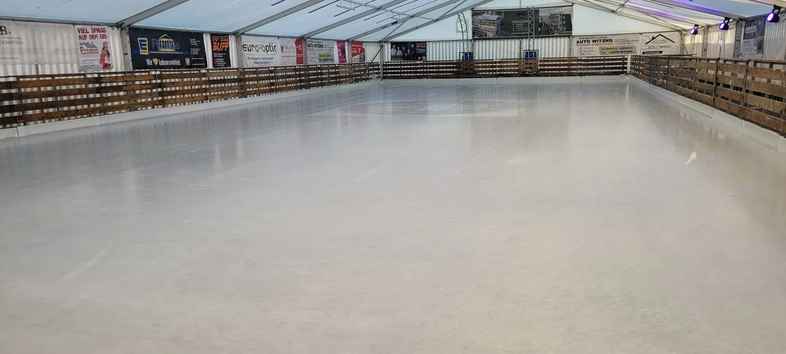 The empty ice surface inside the tent, bounded by low timber boards with sponsor banners along the far side. — Ice4Moor Eisbahn Bad Schwalbach