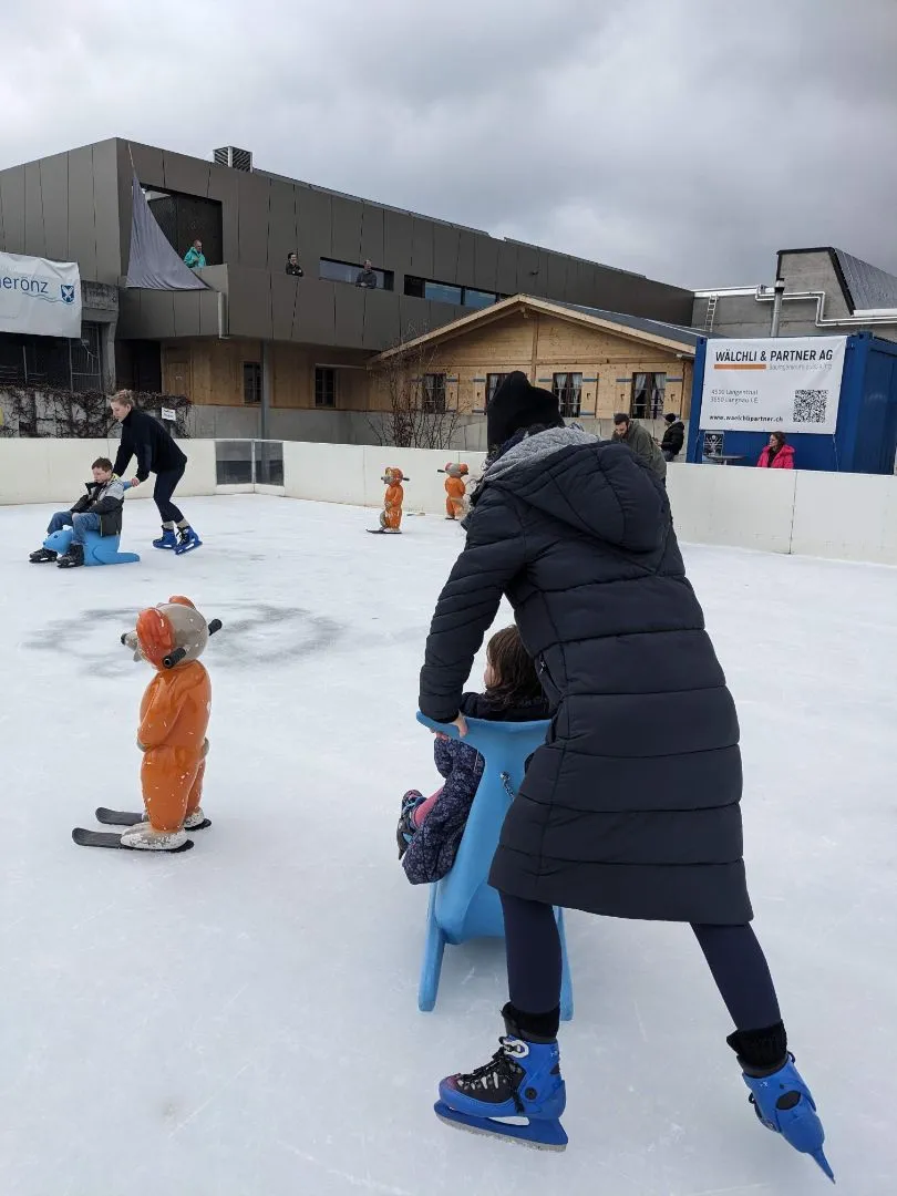 The IceArenA's temporary rink beside the AquArenA building: white boards around the ice, skaters with push-along aids in front, and the grey facade and timber chalet of the AquArenA behind. – IceArenA Herzogenbuchsee