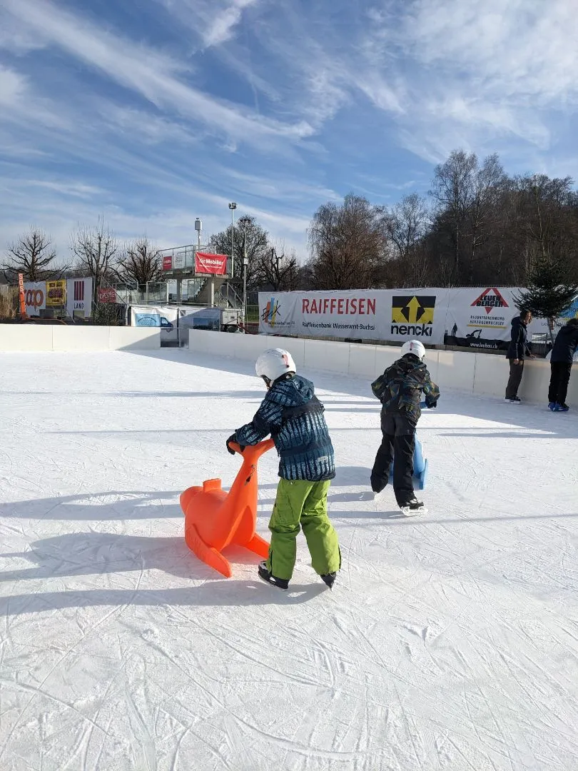 Two children in helmets skating on the white temporary ice, one pushing an orange seal skating aid, with boards, sponsor banners and bare trees beyond. – IceArenA Herzogenbuchsee