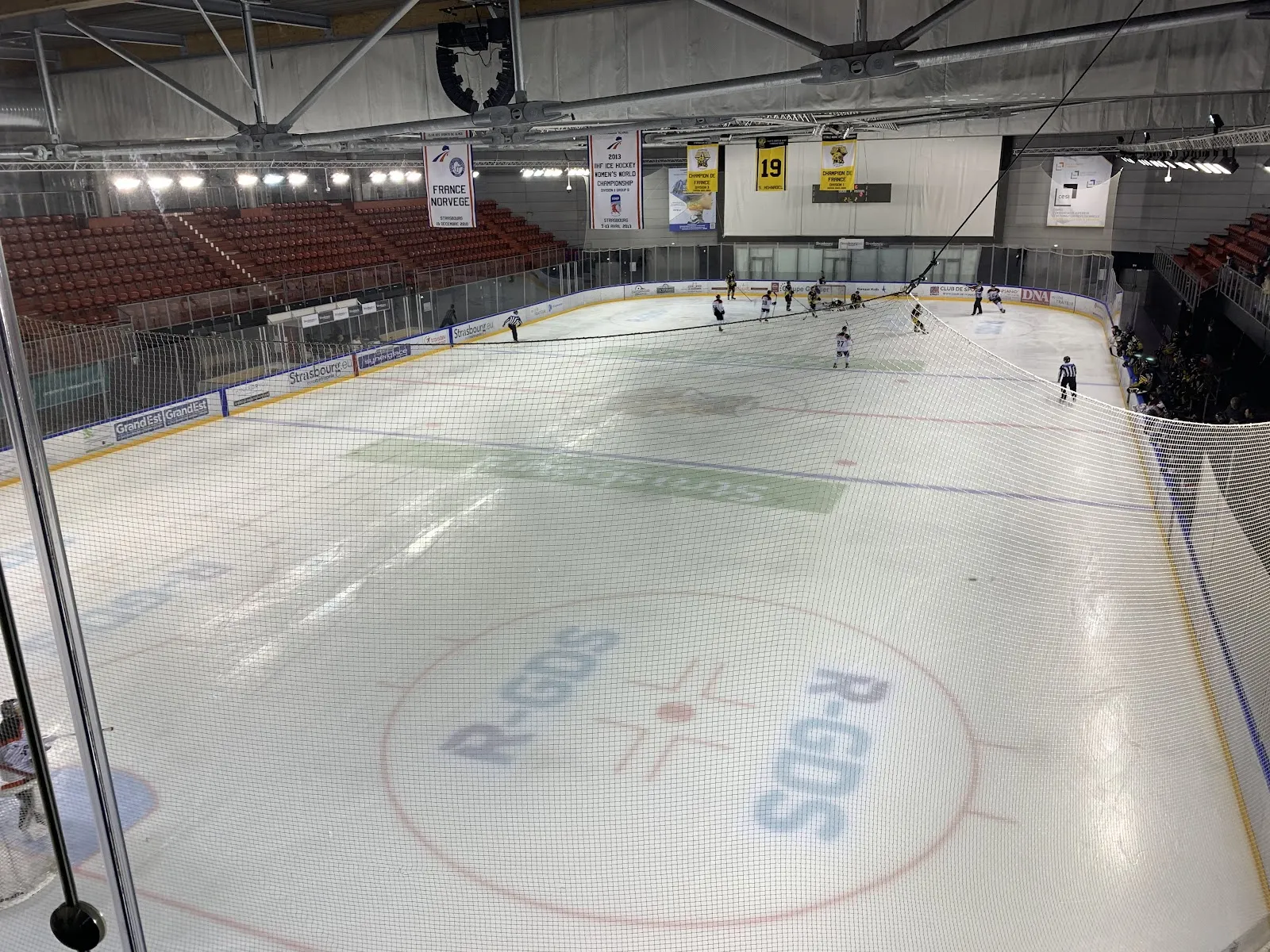 La glace de la piste sportive vue depuis les gradins, avec quelques joueurs au fond et les tribunes couvertes derrière — Patinoire l'Iceberg