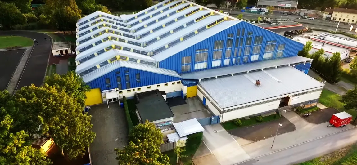 Das Icedome Schweinfurt von oben: eine blau-weiße Halle mit gezacktem Dach, daneben ein Parkplatz und rundherum Bäume – Icedome Schweinfurt