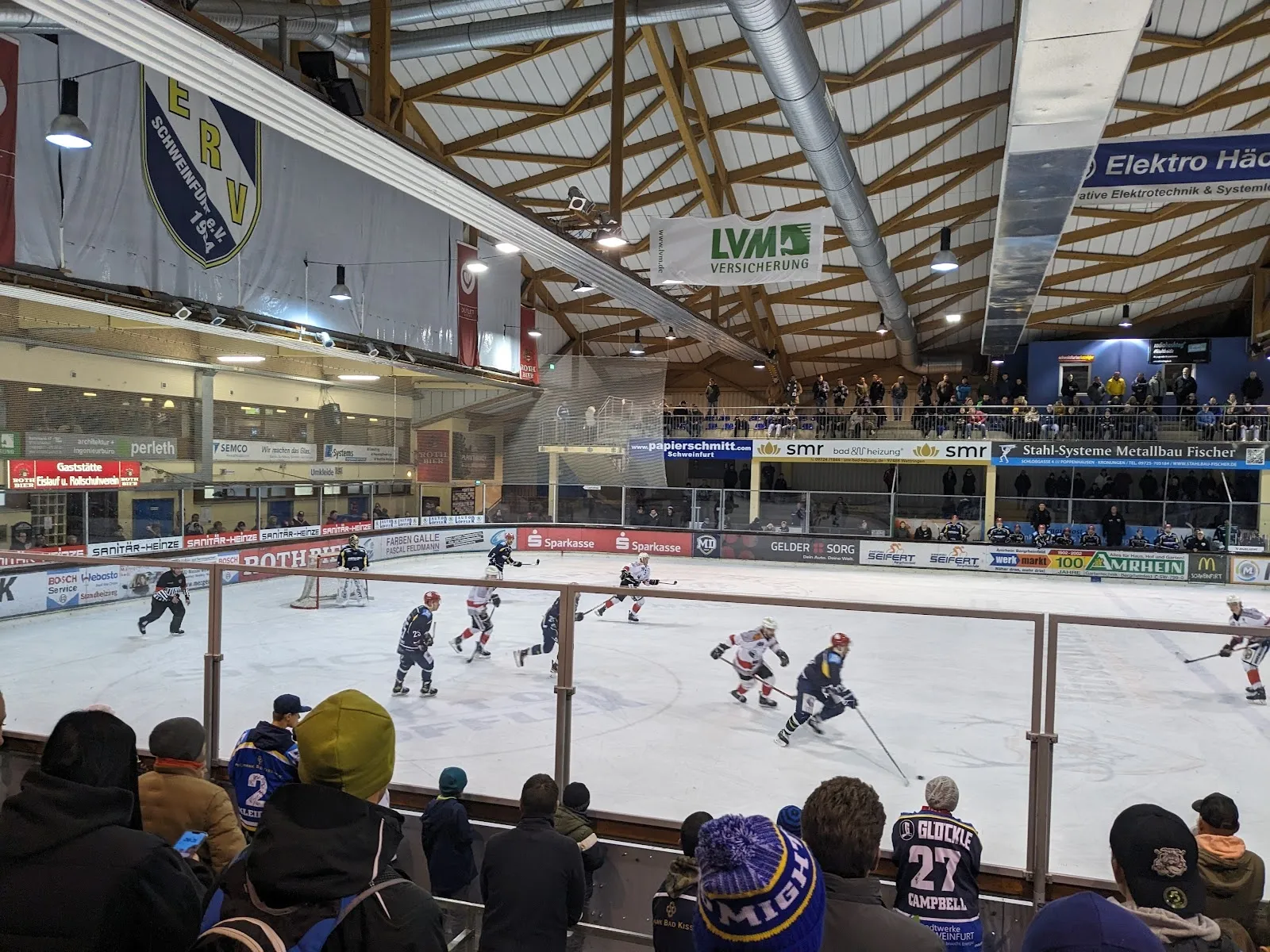 Die Eisfläche des Icedome Schweinfurt während eines Spiels, von den Rängen aus gesehen, mit den Zuschauern rund um die Bande und den Sponsorenbannern darüber. – Icedome Schweinfurt