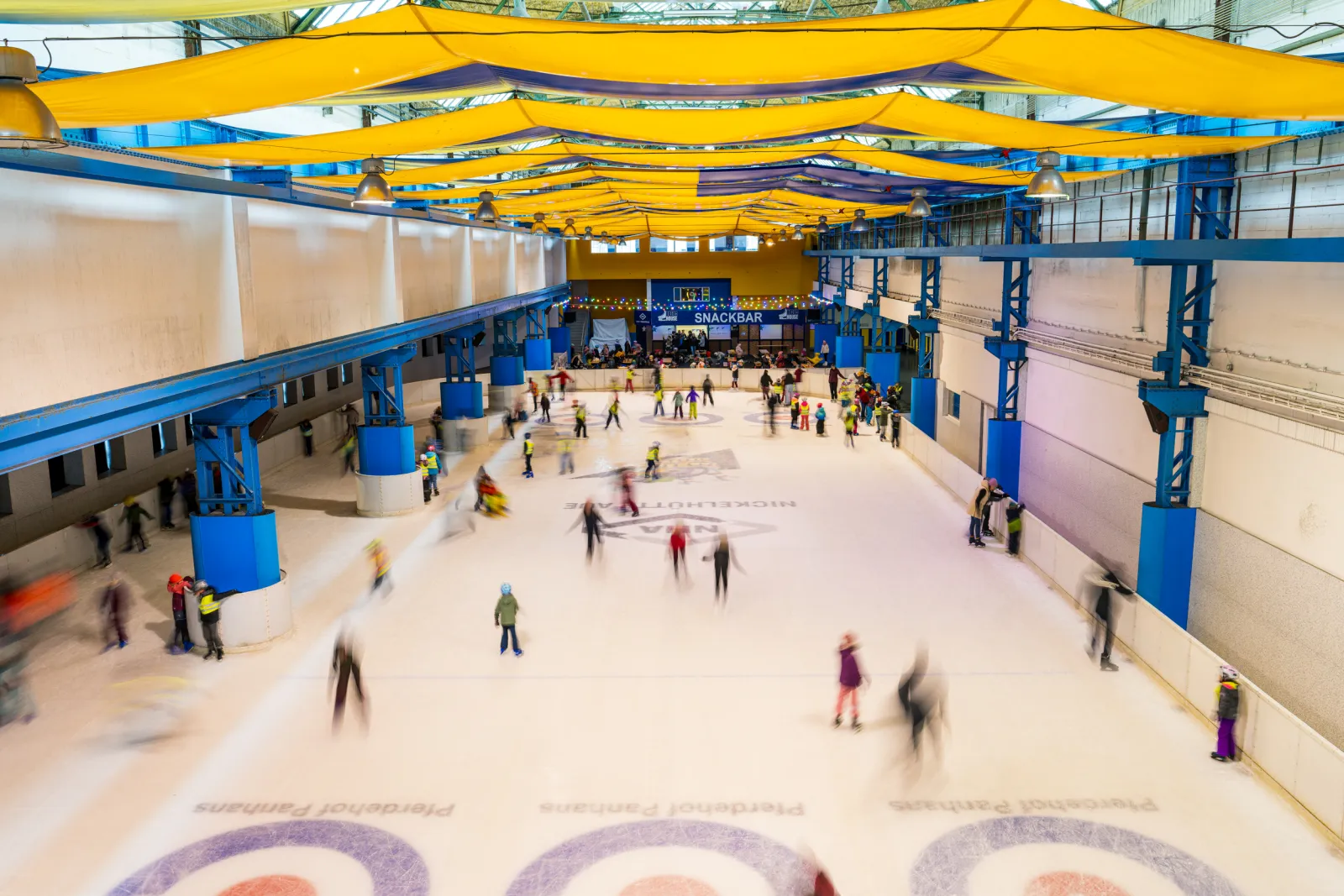 The ice hall of the Icehouse Aue seen from above, the long ice surface with blue steel columns and yellow ceiling sails and skaters spread across it — Icehouse Aue