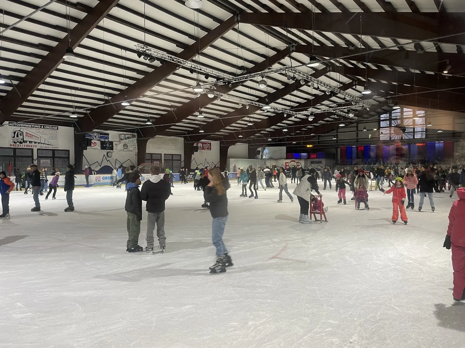 A public skating session on the ice of the Icehouse Eppelheim, with skaters spread across the sheet under the dark timber roof beams and the lighting rig. — Icehouse Eppelheim