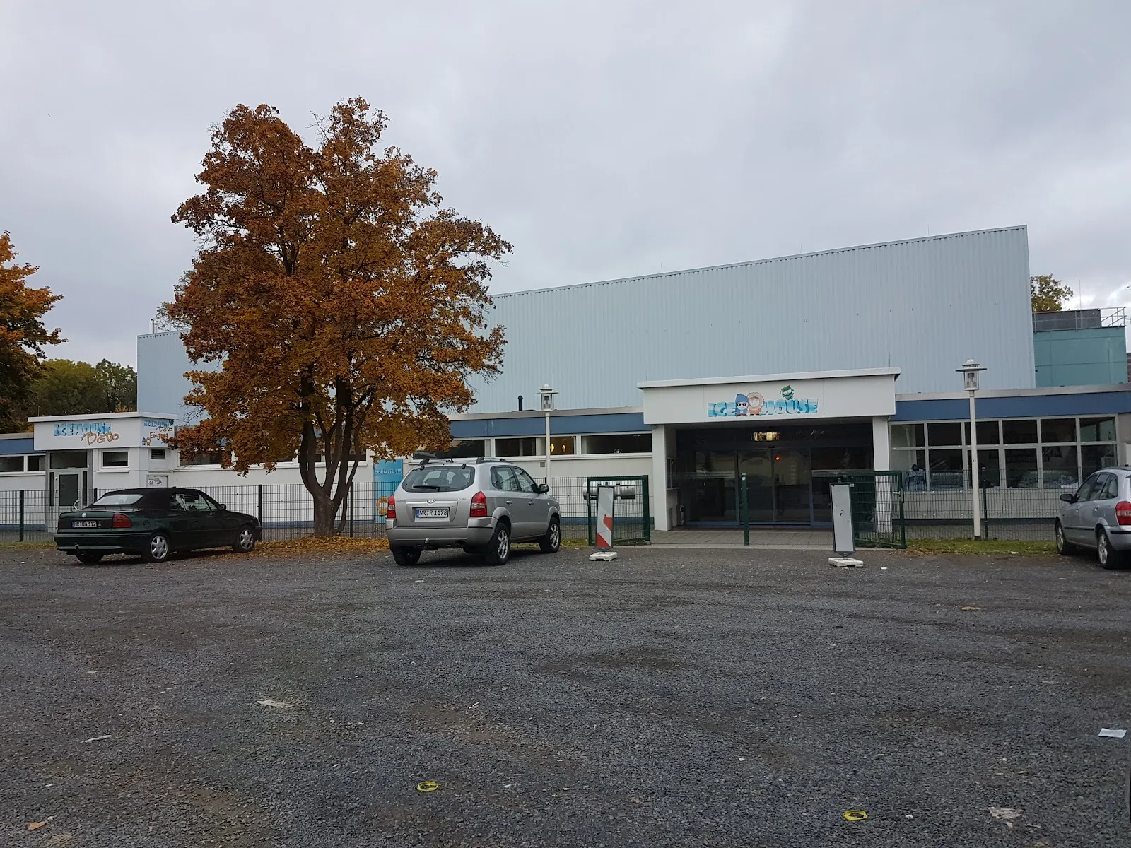 The entrance side of the ice hall behind its gravel car park, with a large autumn tree in front and a grey metal-clad hall roof behind. — Icehouse Neuwied