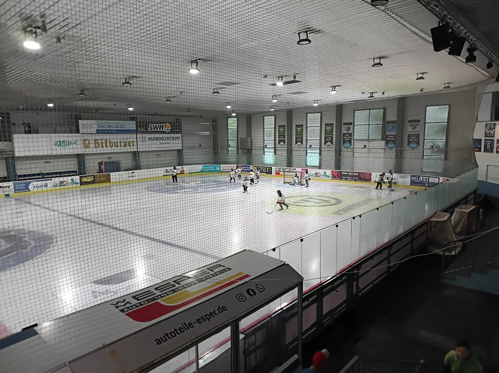 The IceHouse Neuwied rink during a game seen from the stands, players on the ice with sponsor boards and the netted hall around it. — Icehouse Neuwied