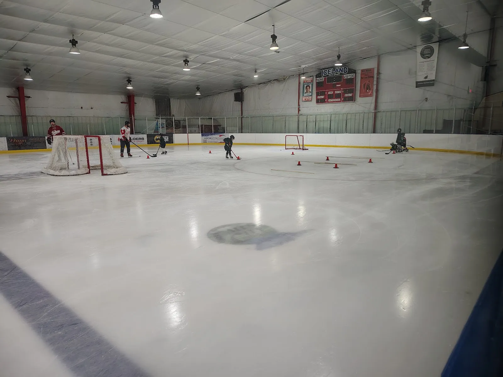 The ice sheet from the glass at one end, with a few skaters near the far boards, a goal at the left and lines under a pale ceiling. — Iceland Sports Complex