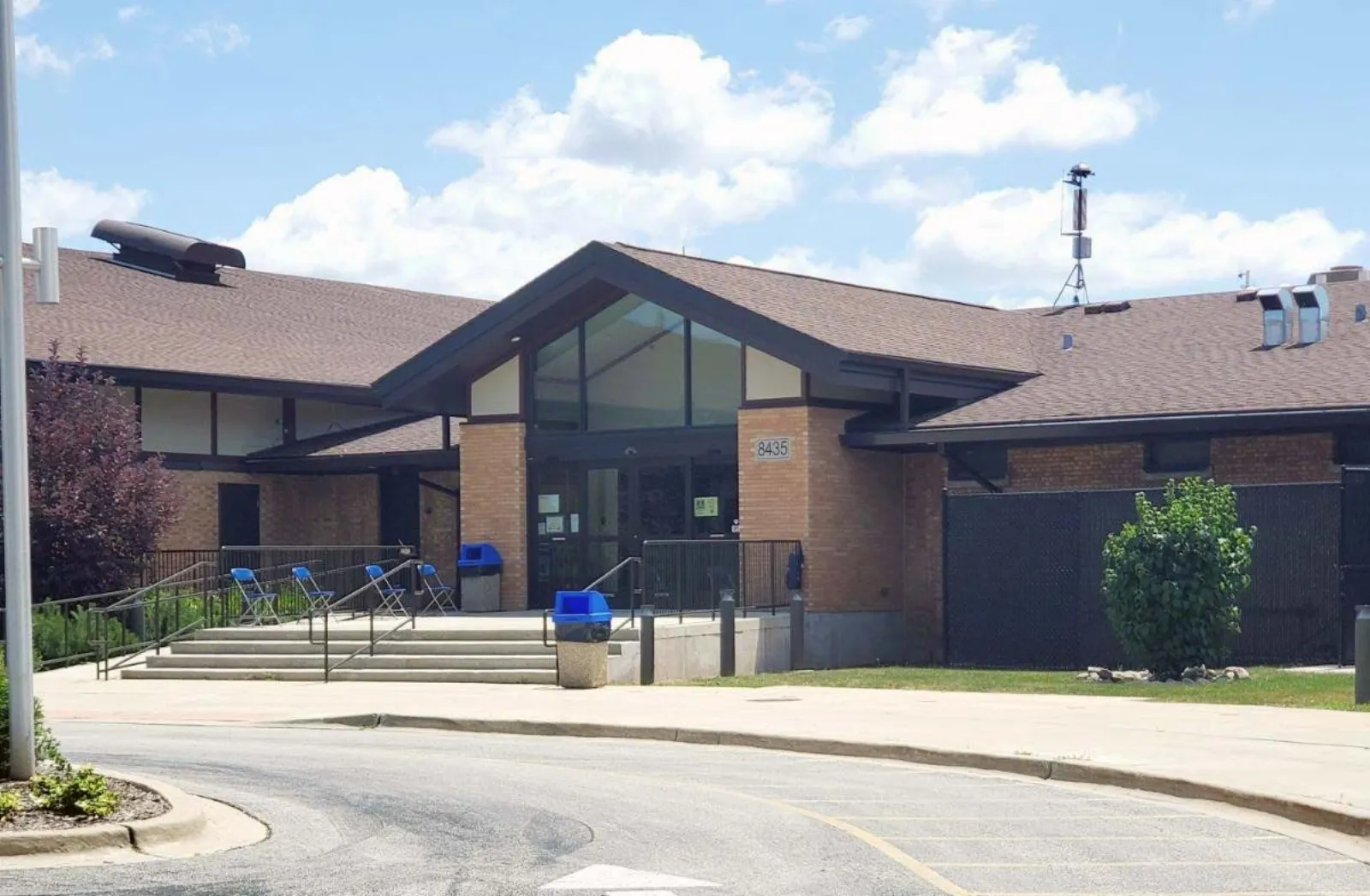 Front entrance of the brick IceLand building with a peaked glass-and-brick entry canopy, the street address "8435" mounted on the brick pillar beside the doors, folding chairs along the entry ramp, and a fenced service area to the right. — IceLand Ice Arena