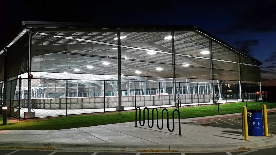 Night view of IceLand's covered outdoor rink structure, a large open-sided metal-roofed pavilion lit by overhead fixtures, with part of the ice sheet and rink boards visible through the chain-link perimeter fence. — IceLand Ice Arena