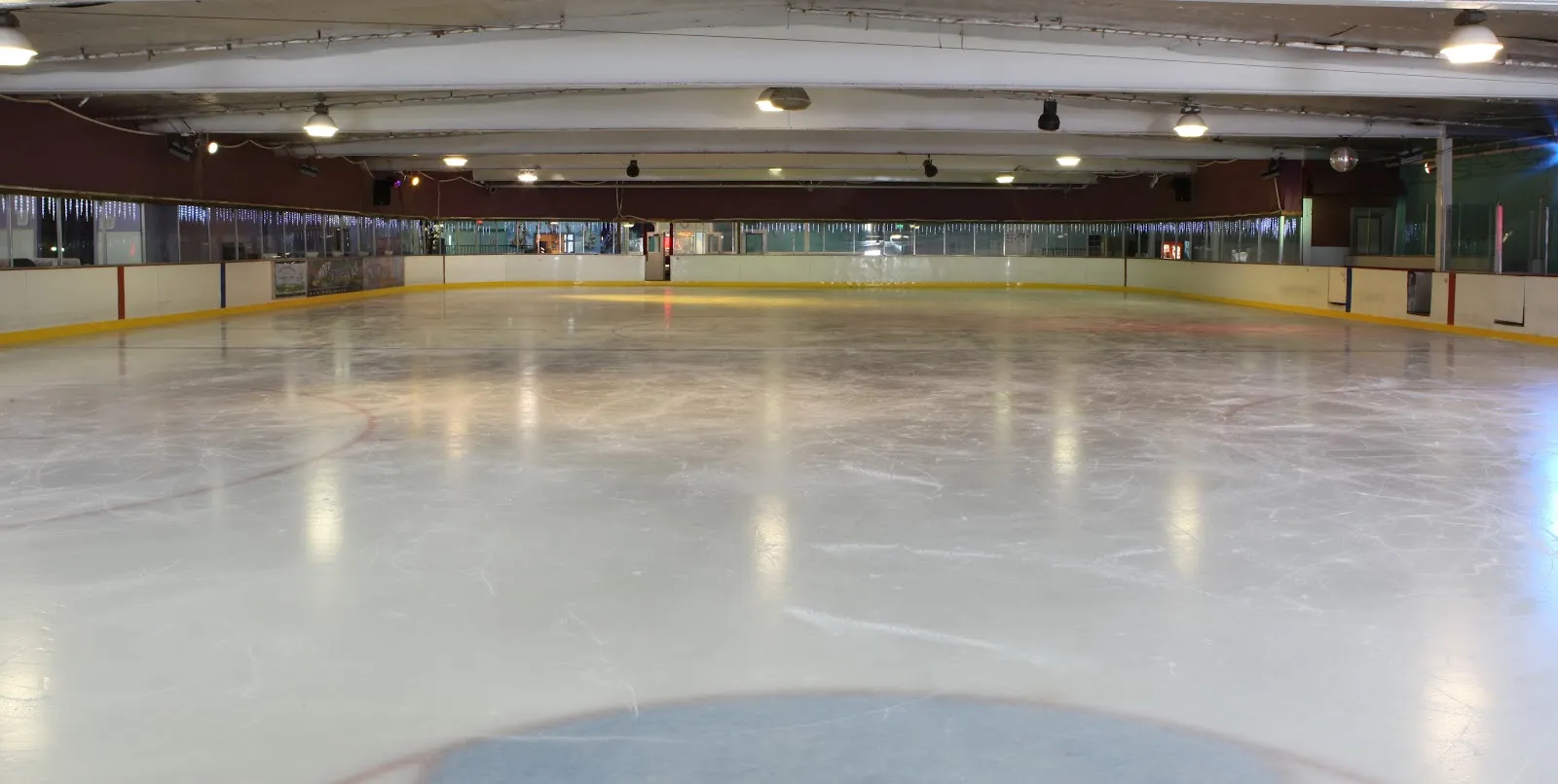 The empty sheet seen along the boards, with a yellow kickplate and the pale low ceiling above. — Iceland Ice Skating Center