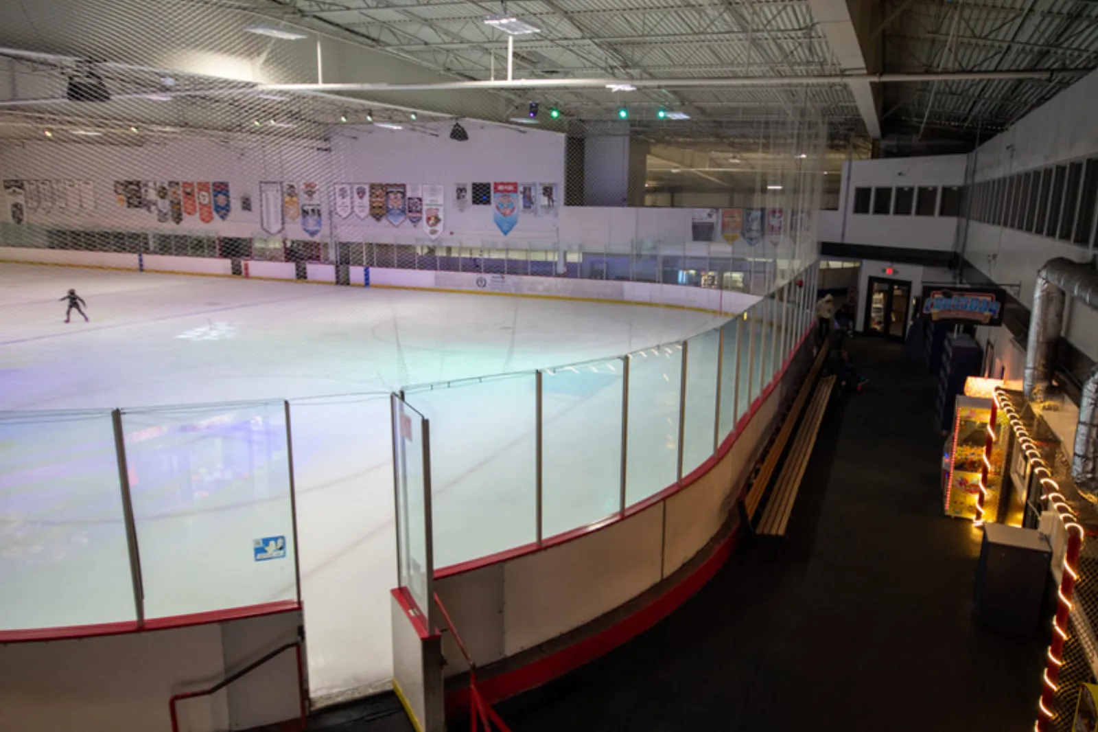 Vista elevada de toda la pista de tamaño NHL desde el pasillo superior, con las vallas, mantas de clubes y ligas en la pared del fondo y un solo patinador artístico practicando en el hielo. — Iceoplex