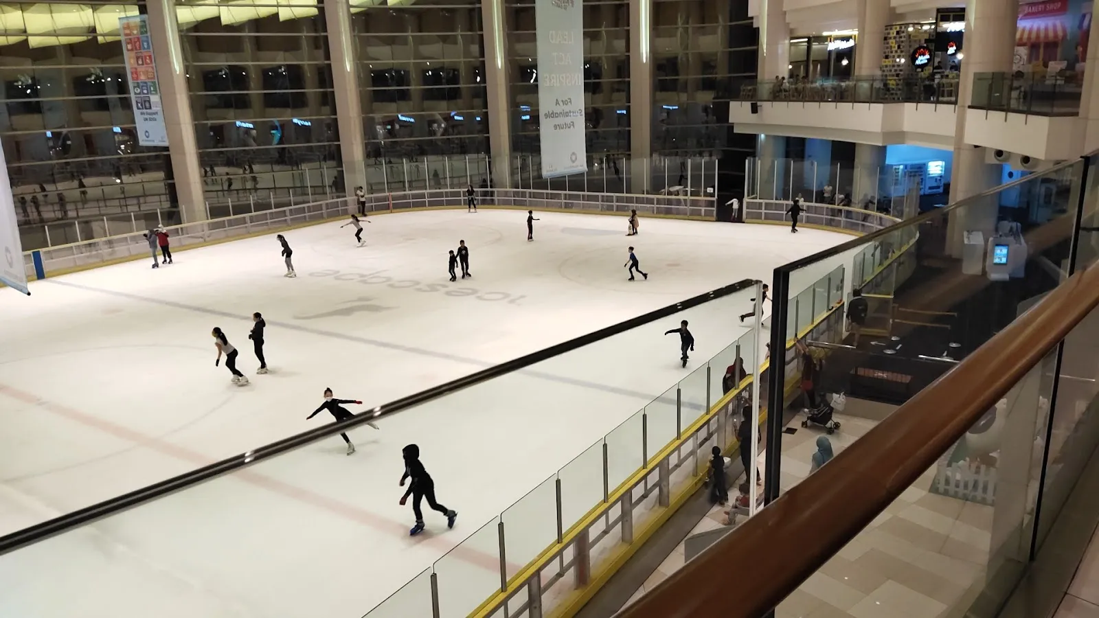 The sheet seen from the mall's upper level, with the glass barrier, the viewing gallery and a few skaters on the ice. — Icescape Ice Rink