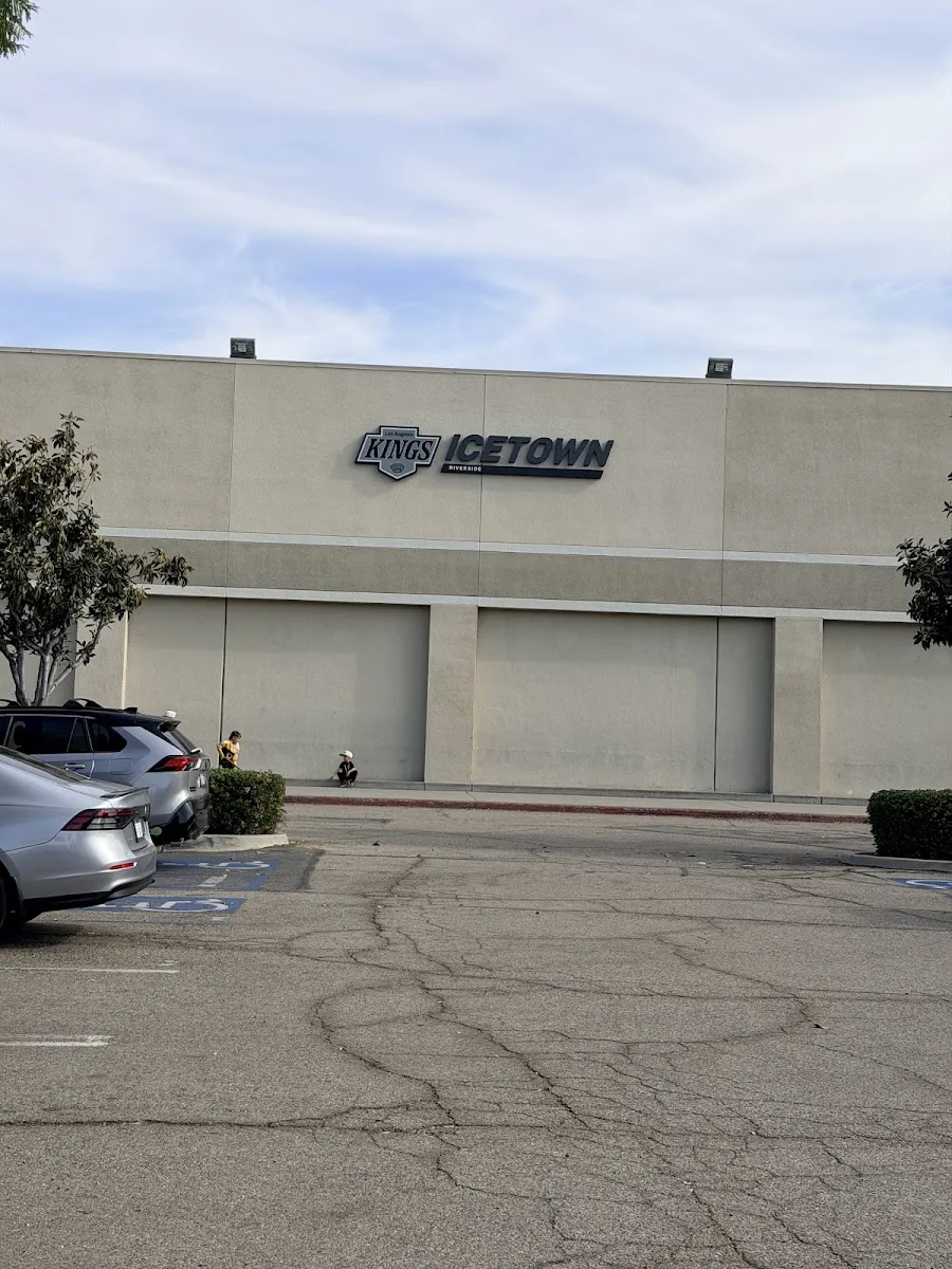 Front of the Icetown Riverside building, tan stucco facade with the LA Kings ICETOWN Riverside wordmark and shield logo mounted above closed roll-up doors, parking lot in front. — Icetown Riverside