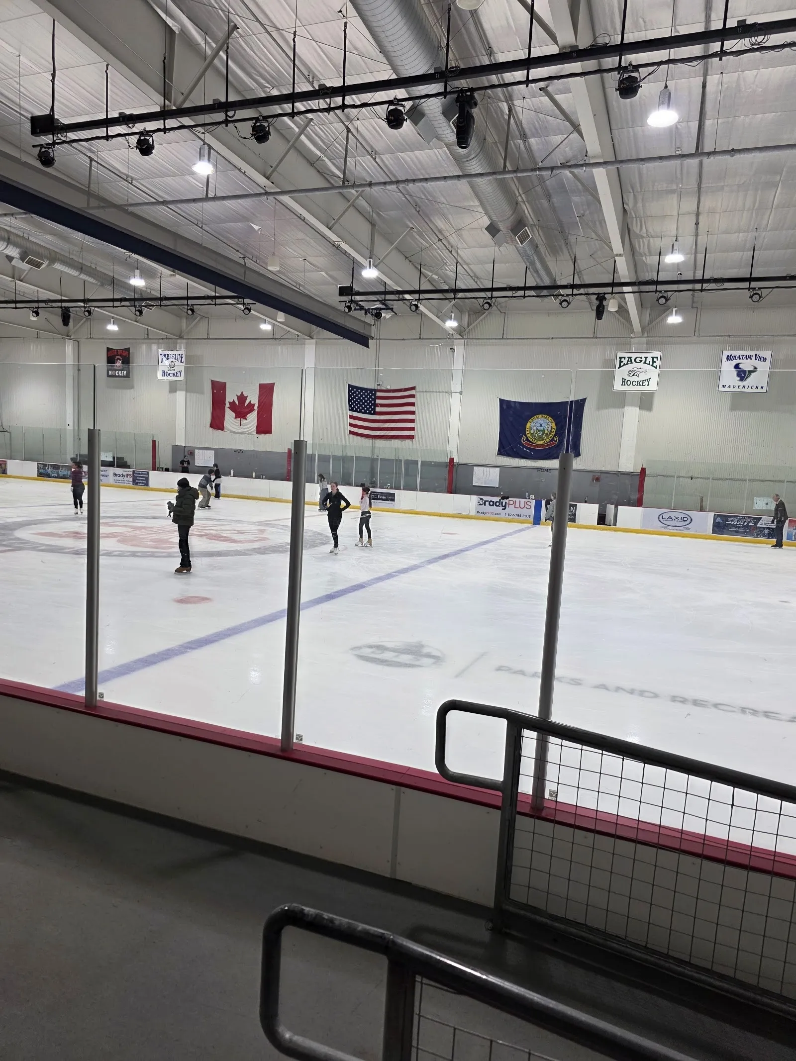 Una sesión pública en una pista amplia vista desde las gradas, con patinadores repartidos por el hielo bajo banderas de Canadá, Estados Unidos e Idaho en la pared del fondo. — Idaho IceWorld