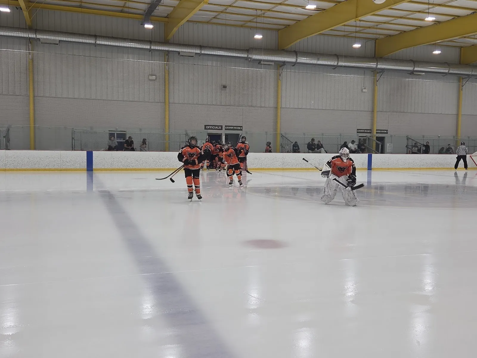 Una vista amplia de la pista con jugadores repartidos por el hielo y un techo de armaduras amarillas, con círculos de saque y líneas azules sobre la superficie blanca. — Igloo Ice Rink