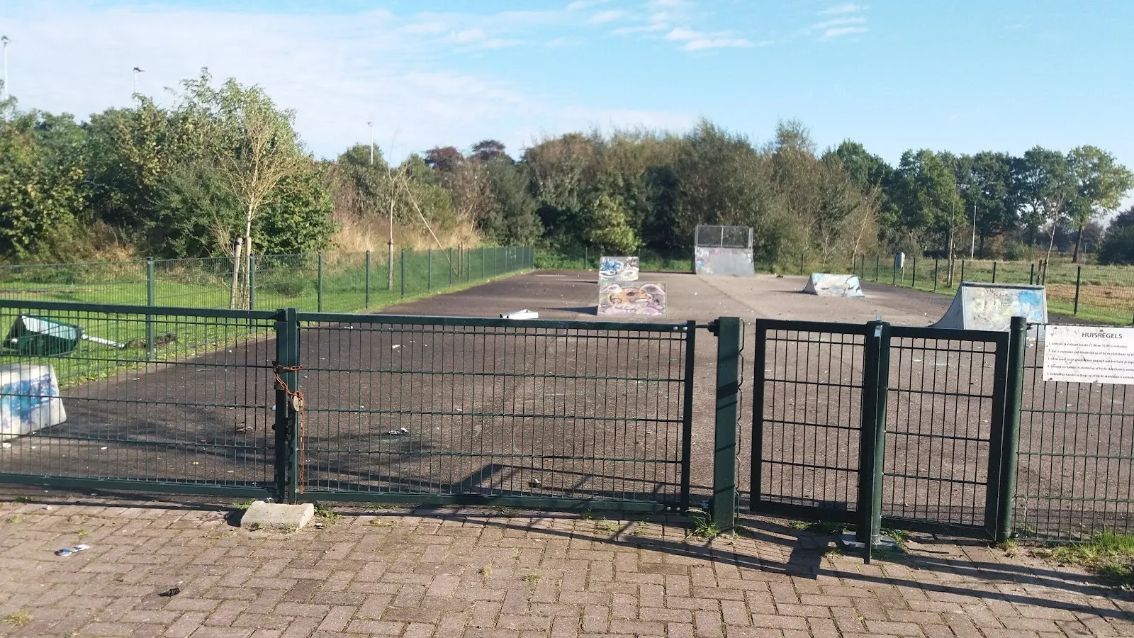 Het hek bij de ingang van het ijs- en skeelerbaangebied in Surhuisterveen op een zonnige herfstdag, met een bestraat voorterrein, bomen en de baan erachter. – Iisclub Foarút (IJsbaan Surhuisterveen)