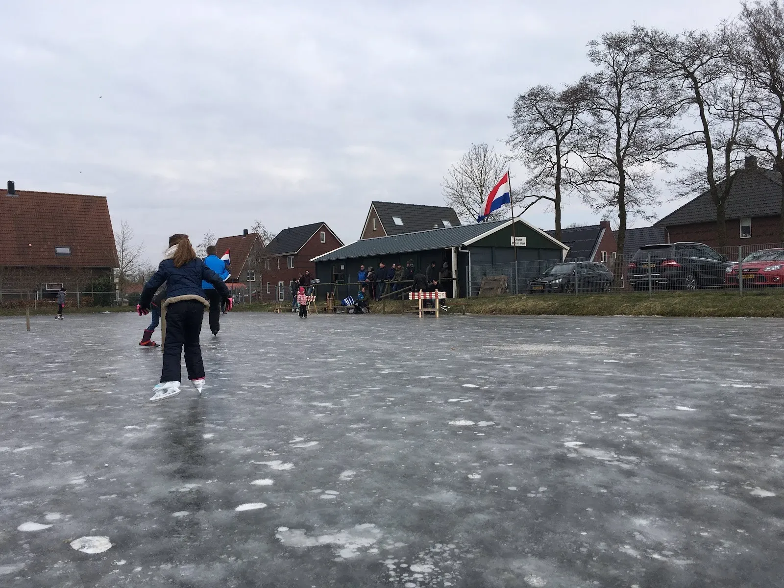 Een schaatser loopt over dun, gespikkeld ijs op de baan in Surhuisterveen, met dorpshuizen, kale bomen, een clubgebouw en een Nederlandse vlag langs de overkant. – Iisclub Foarút (IJsbaan Surhuisterveen)