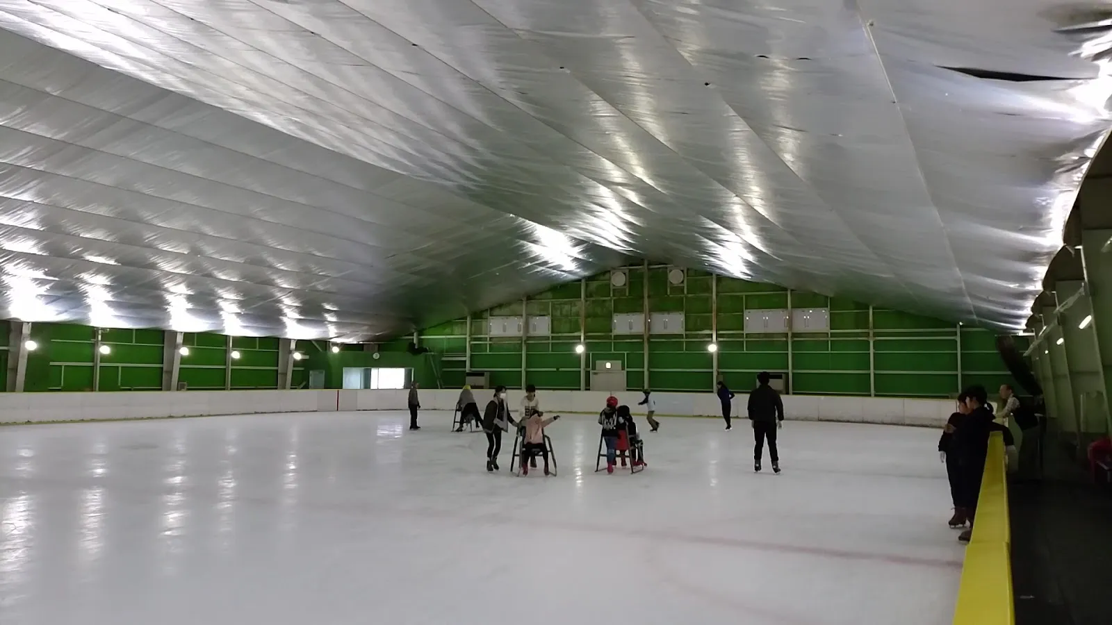 The wide ice sheet of Iizuka Ice Palace under its silver foil-lined ceiling, with a handful of skaters in the middle distance and green boards at the far end. — Iizuka Ice Palace