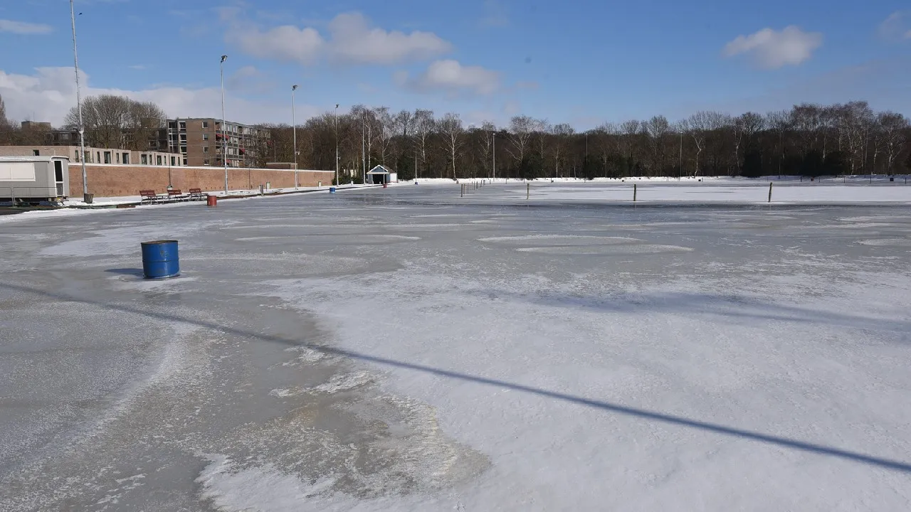 De natuurijsbaan van Wolvega in de winter, met gebarsten ijs en sneeuw, lichtmasten, een klein huisje en bankjes aan de overkant en kale bomen erachter. – IJsbaan Wolvega (IJs- en Skeelerclub Lindenoord)