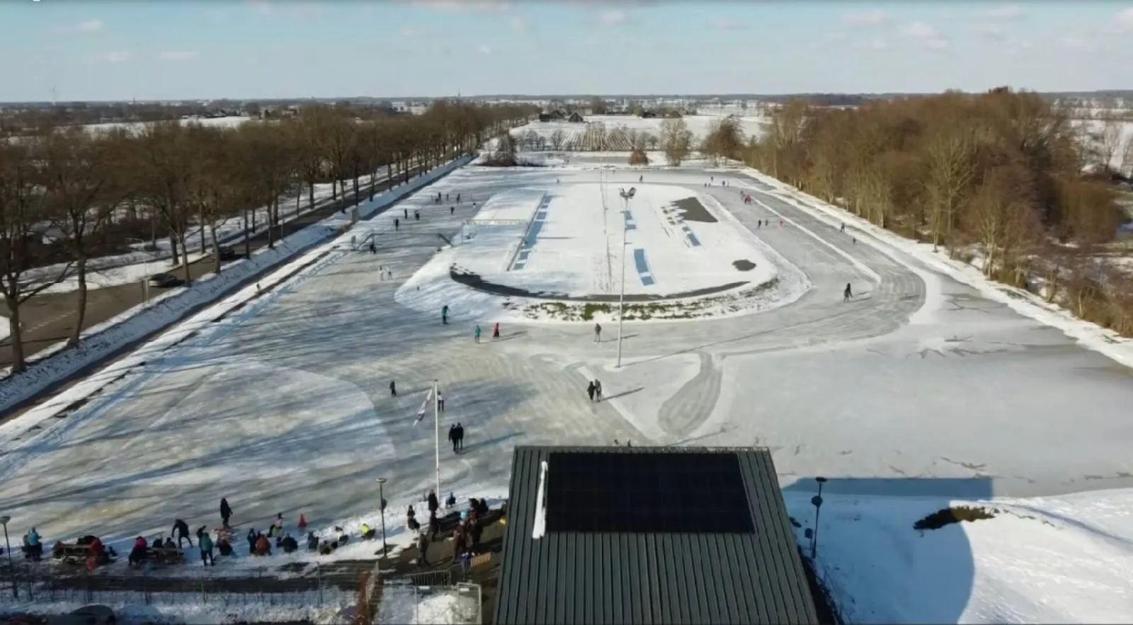 An aerial view of the snow-covered natural-ice track at Nijeveen with skaters along the edge and a roof in the foreground, long avenues of trees running beside the rink. — IJs- en Skeelerclub Nijeveen