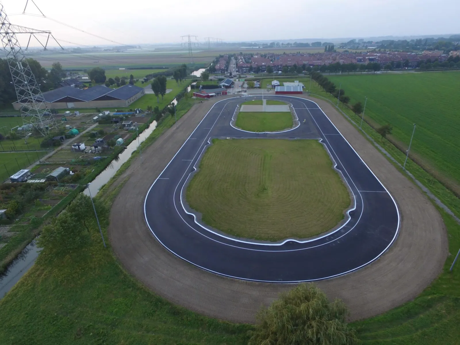 Aerial view of the club's 200 m and 400 m asphalt tracks at Puttershoek, the oval with a grass infield, with farmland, a ditch and the village behind. — IJsbaan Puttershoek (IJs- en Skeelerclub Puttershoek)