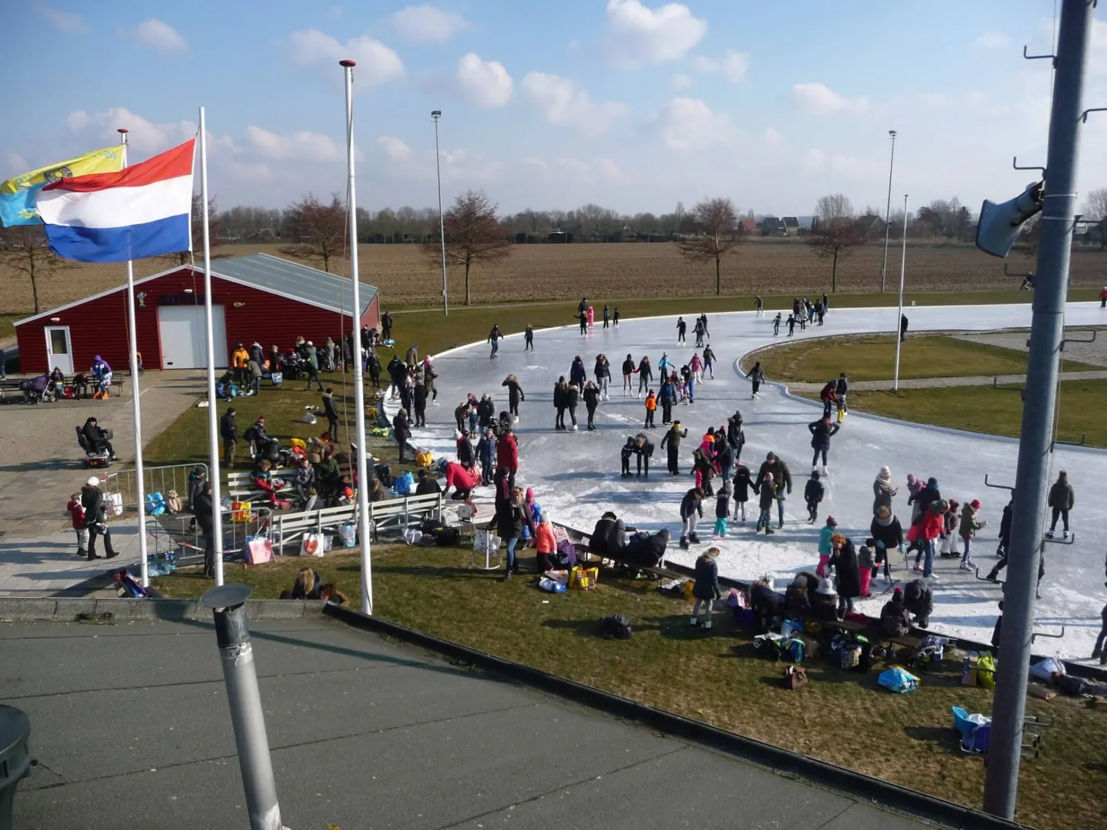 Many skaters and families on the pale ice of the club's oval track at Puttershoek on a bright day, with the red clubhouse, flagpoles and floodlight masts beside it. — IJsbaan Puttershoek (IJs- en Skeelerclub Puttershoek)