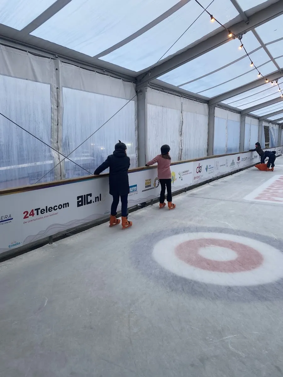 Two children seen from behind at the boards of the covered ice rink of IJs in Ermelo, with the curling-style ring painted on the ice — IJs in Ermelo