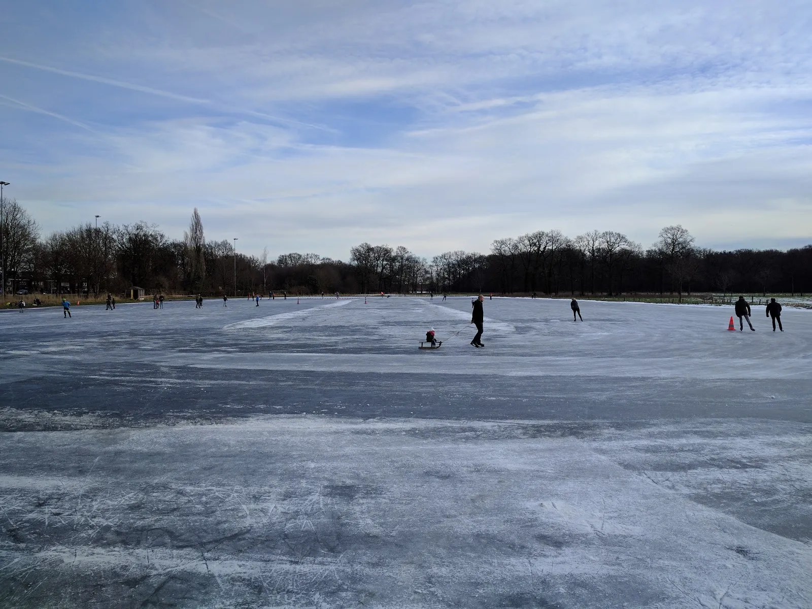 Schaatsers ver uit elkaar op het brede natuurijs van de Almelose ijsbaan onder een winterse lucht, met lichtmasten en bomen langs de verre rand. – IJsbaan Almelo