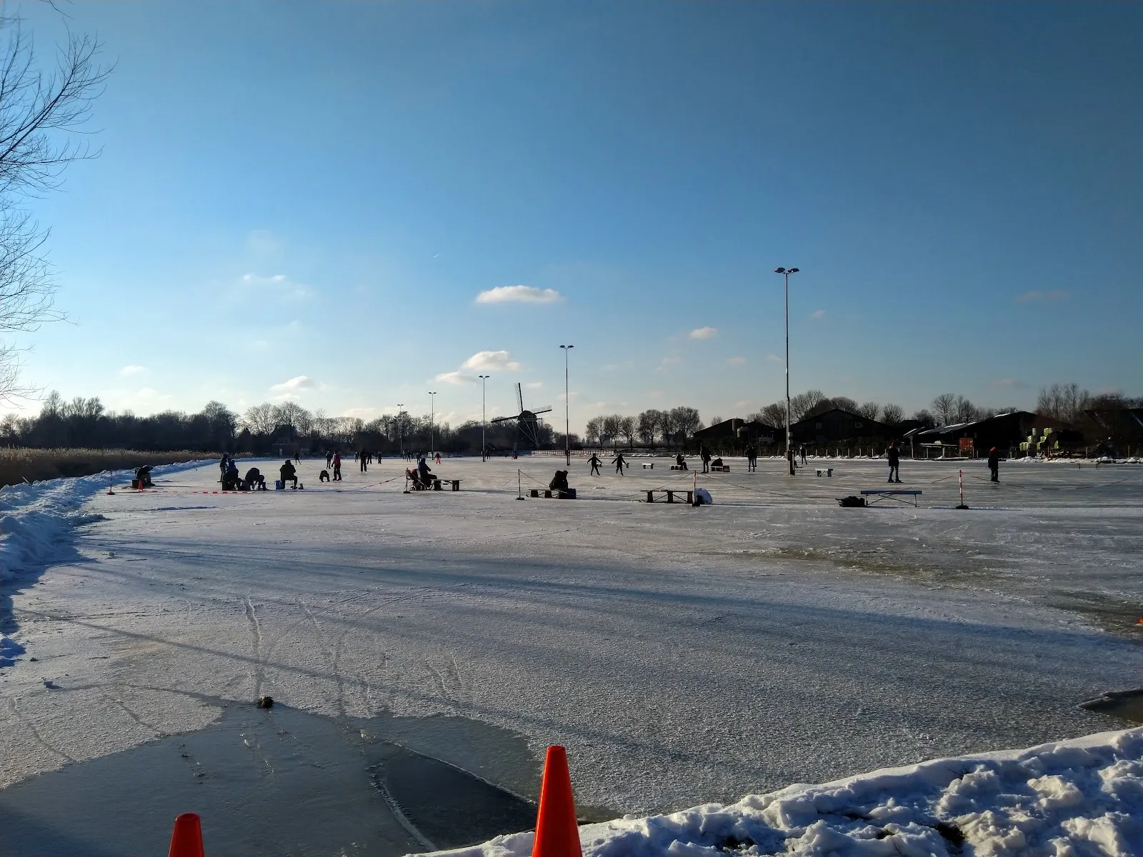 Natural ice on the Amstelbocht rink under a blue winter sky, with skaters spread out in the distance and snow along the edge. – Landijsbaan De Amstelbocht (Ouderkerk aan de Amstel)