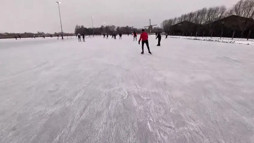 Skaters crossing the smooth ice of the Amstelbocht rink on a bright day, with the dyke and trees behind. – Landijsbaan De Amstelbocht (Ouderkerk aan de Amstel)