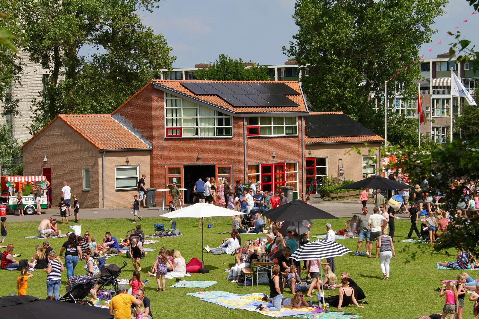 Het rode bakstenen clubhuis van IJsclub Voorwaarts op sportpark De Goerie op een zomerse dag, met bezoekers op het gras ervoor onder parasols. – Landijsbaan De Goerie (Katwijk)
