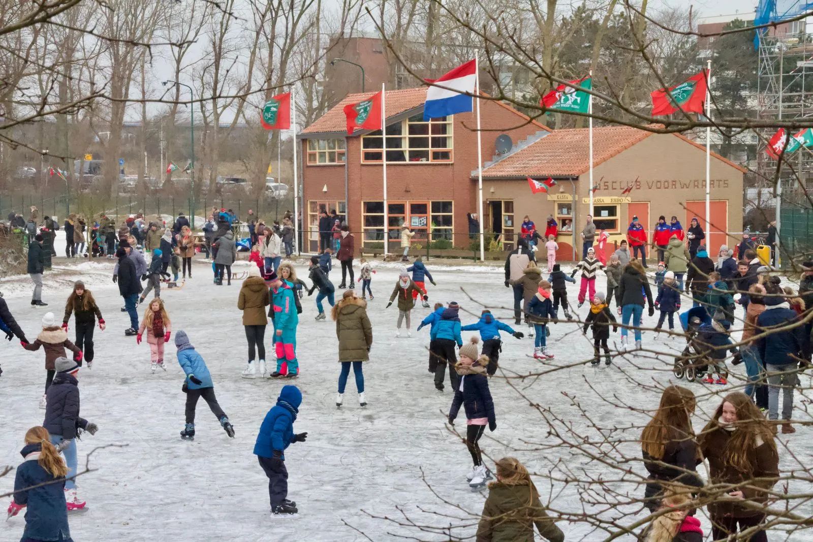 Een menigte gezinnen schaatst op het dunne ijs van de landijsbaan De Goerie voor het rode bakstenen clubhuis, met wapperende vlaggen en kale bomen erachter. – Landijsbaan De Goerie (Katwijk)