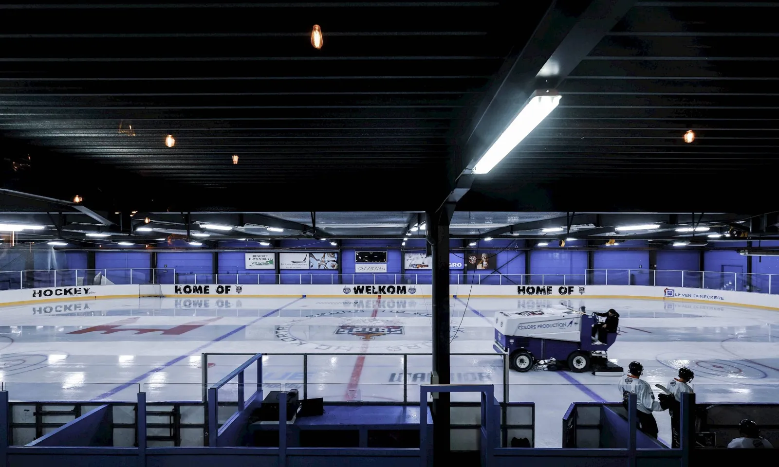 De overdekte ijsbaan van Ijsbaan de Piste in Kortrijk onder een donker dak met tl-balken, een paarse hockeyband eromheen en een ijsmachine aan het werk met twee medewerkers ernaast. – IJsbaan de Piste (Kortrijk)