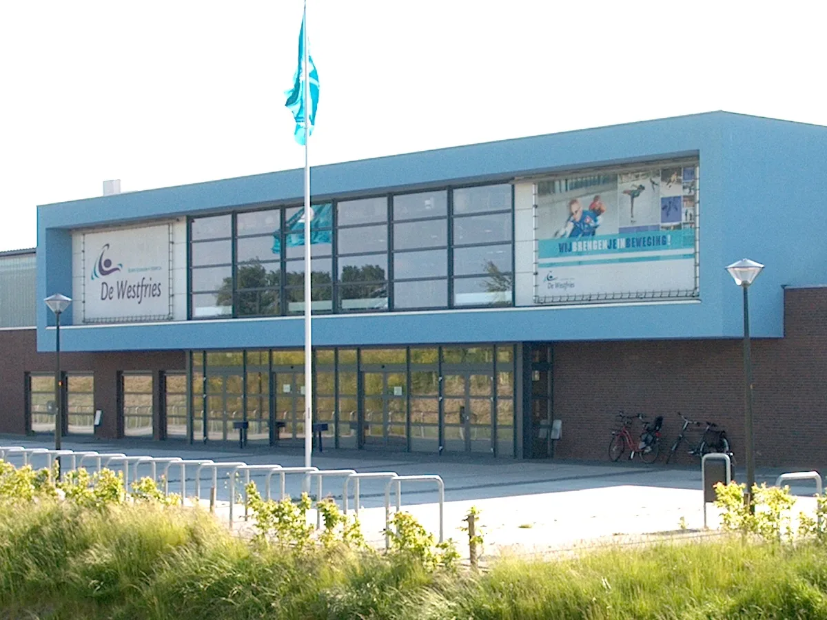 The entrance side of IJsbaan De Westfries in Hoorn, a blue and brick building with the De Westfries name sign and a flagpole in front of the glass entrance — IJsbaan De Westfries