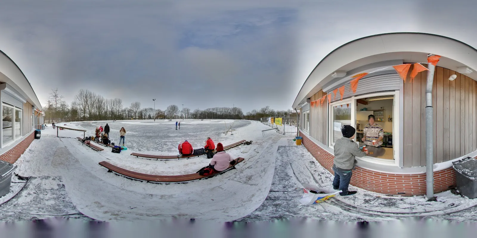 Het clubhuis met zijn uitgifteluik en oranje vlaggetjes naast het met sneeuw bestoven ijs, met banken langs de rand. – IJsbaan Dicky van der Werf (IJsclub Sneek)